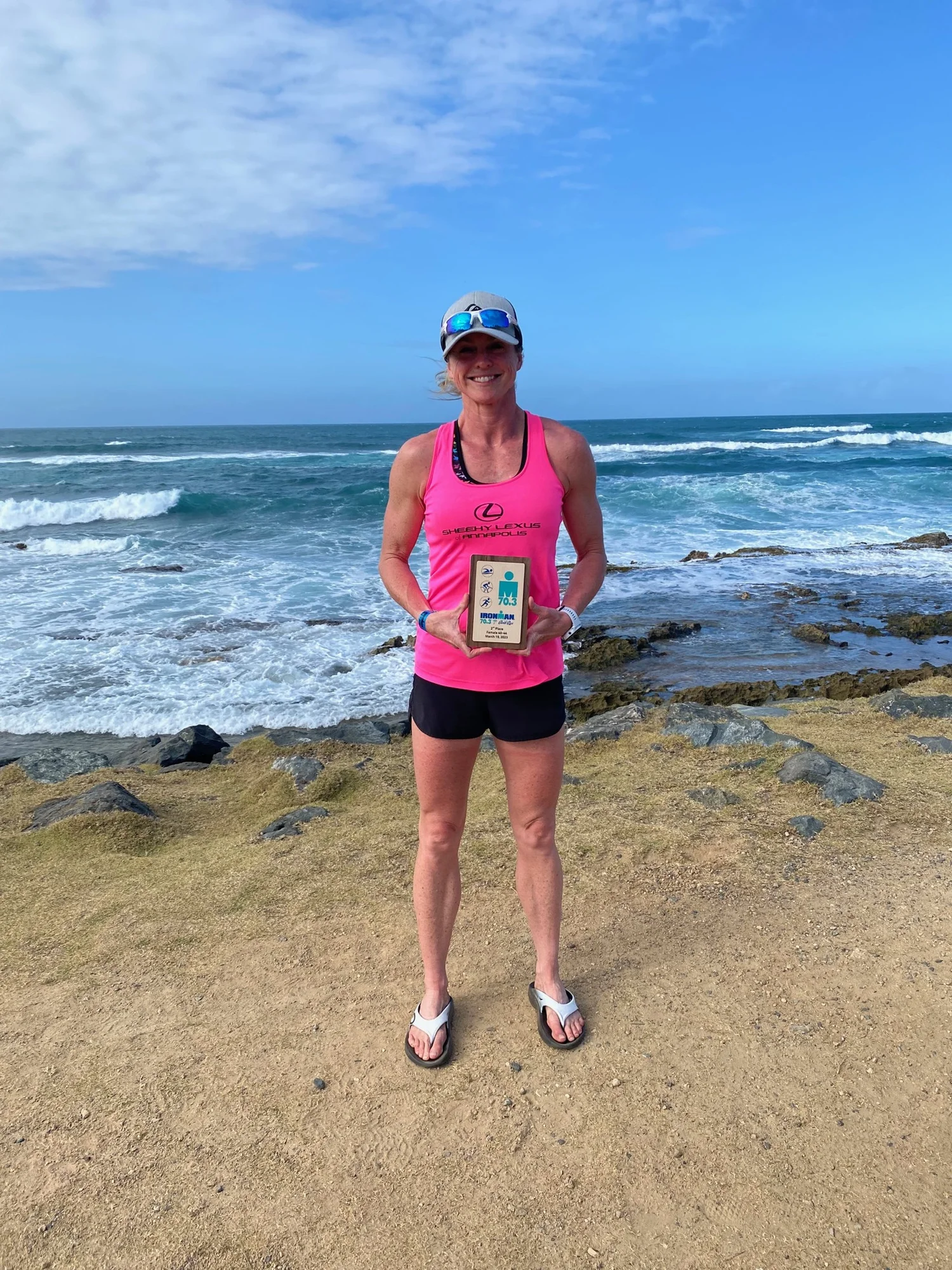 A woman in a pink tank top and black shorts stands on a sandy beach near the ocean, holding an award plaque from an IRONMAN Triathlon.