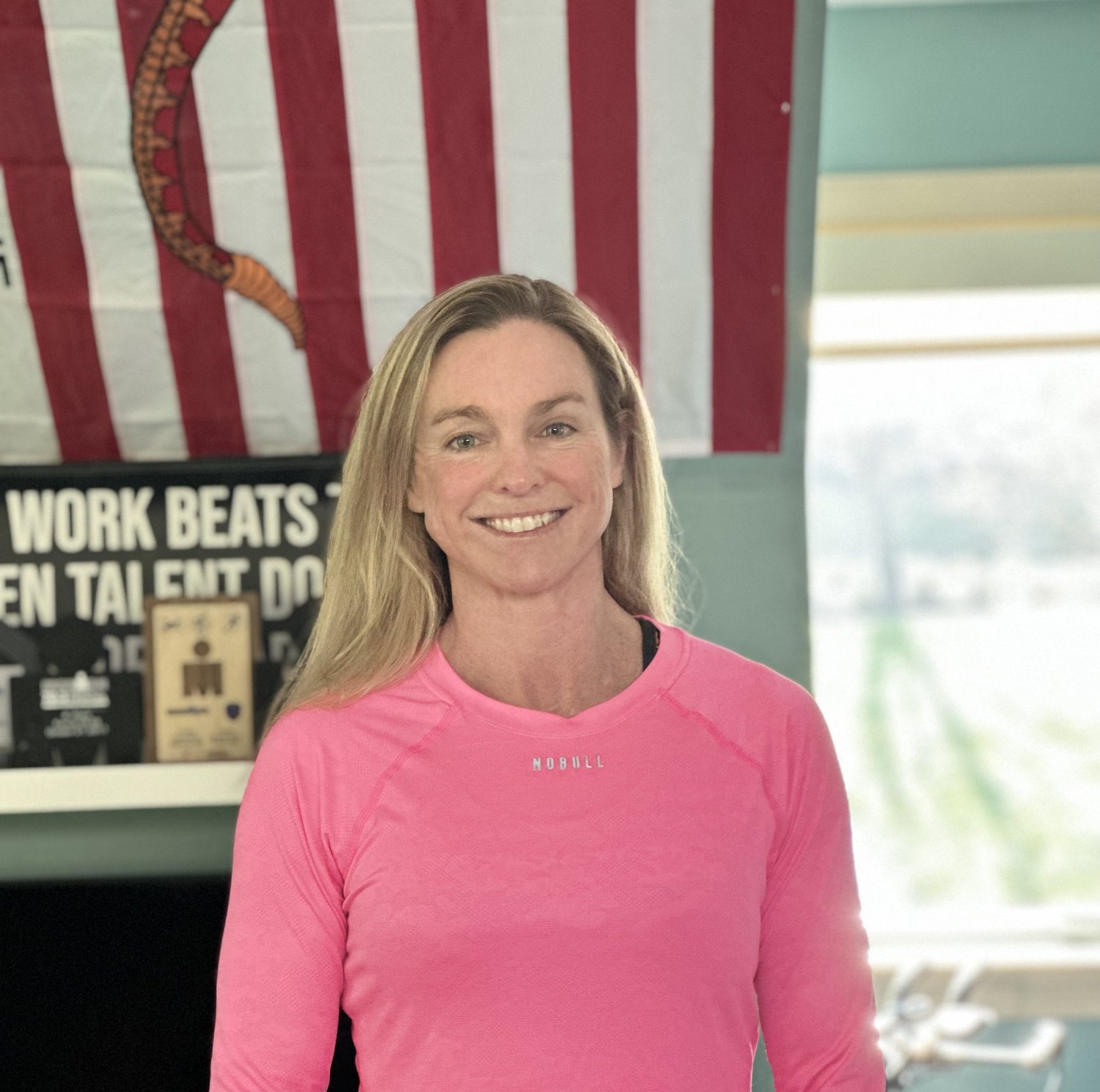 Alli LaRochelle with long blonde hair smiling, wearing a pink athletic shirt indoors.