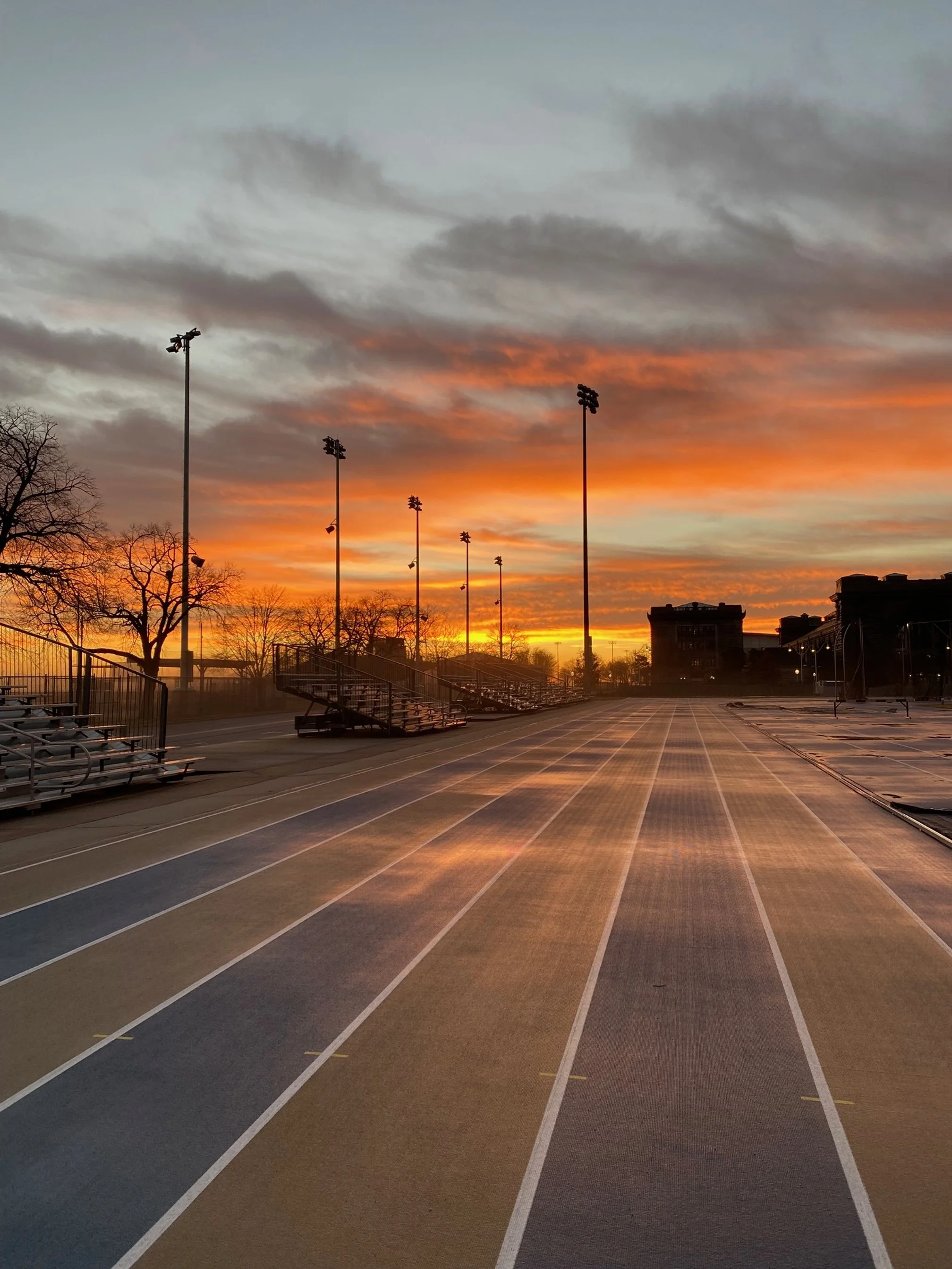 Empty outdoor track and bleachers at sunset with colorful sky and scattered clouds.