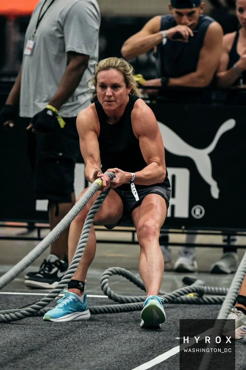 A woman competing at a HYROX event, pulling a sled in athletic gear inside an indoor gym.