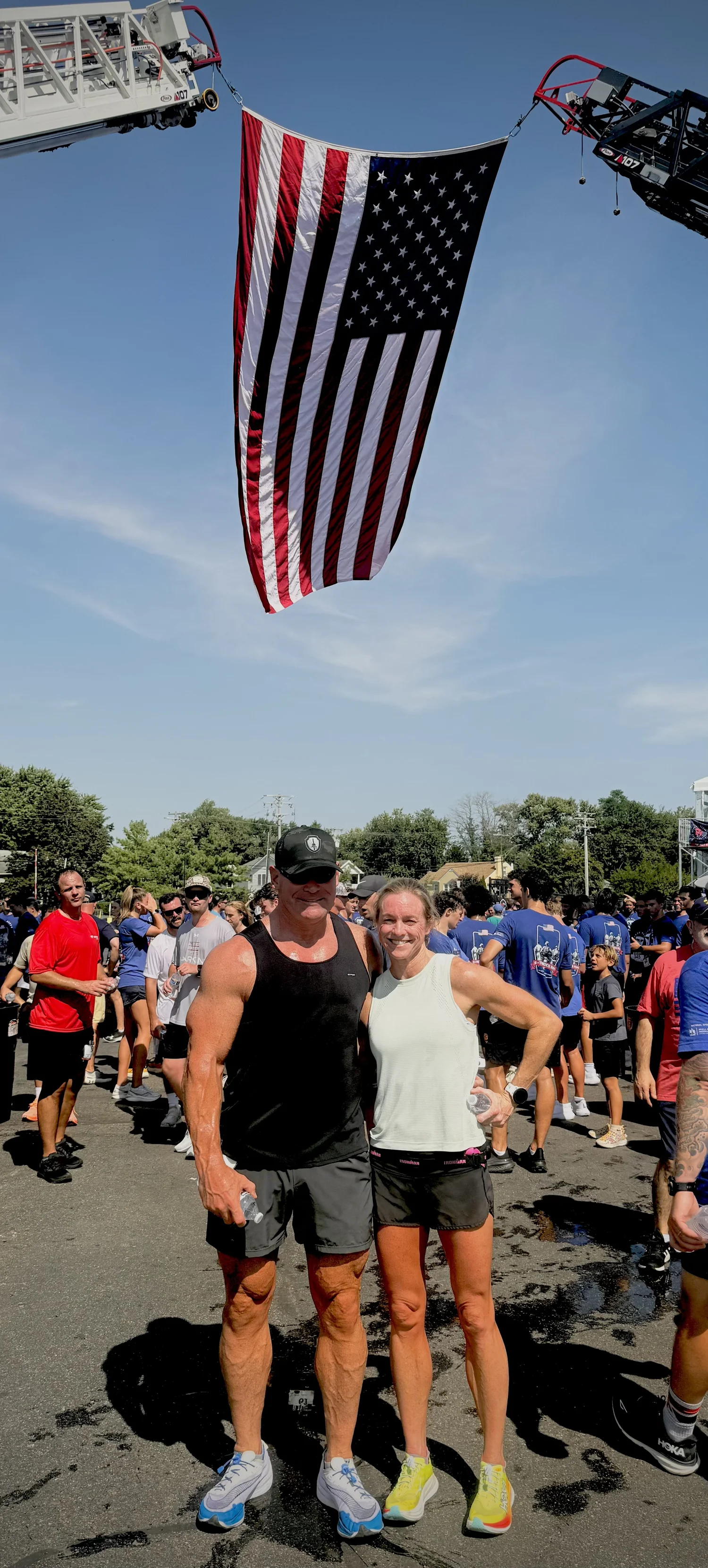 A large American flag hanging between two fire trucks during a public event, with two smiling athletes in the foreground.